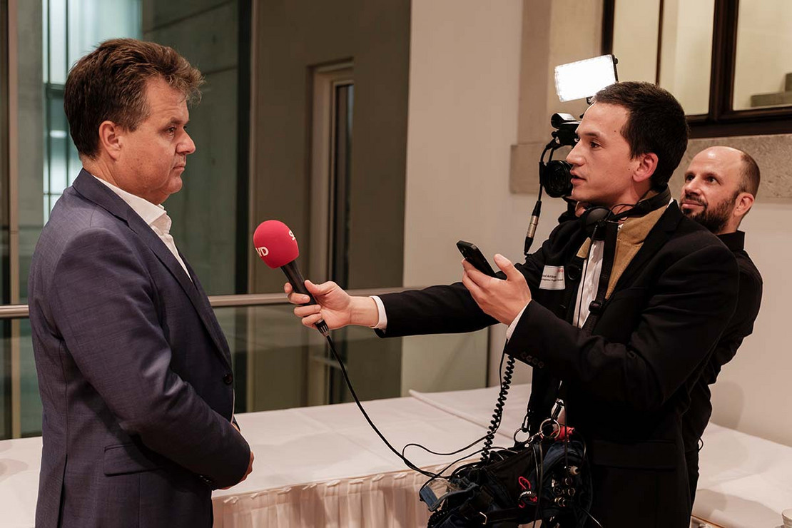 Mann spricht in ein rotes SoVD-Mikrofon und wird dabei von einem Kamerateam gefilmt.