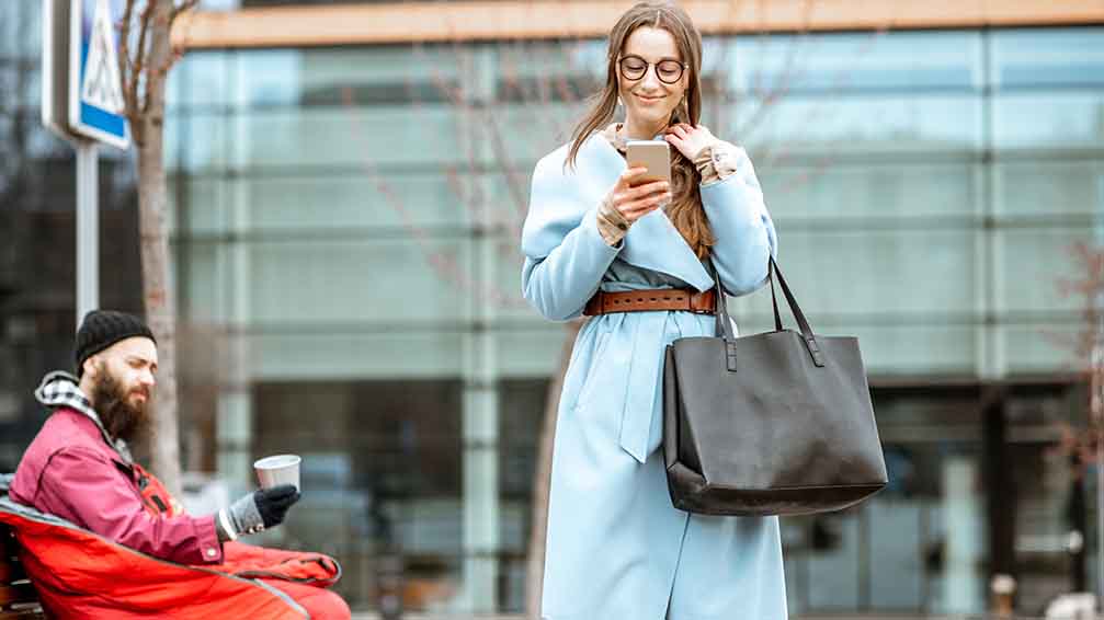 Elegante Frau mit Smartphone läuft an einer Bank vorbei. Darauf sitzt ein Mann und bettelt. 