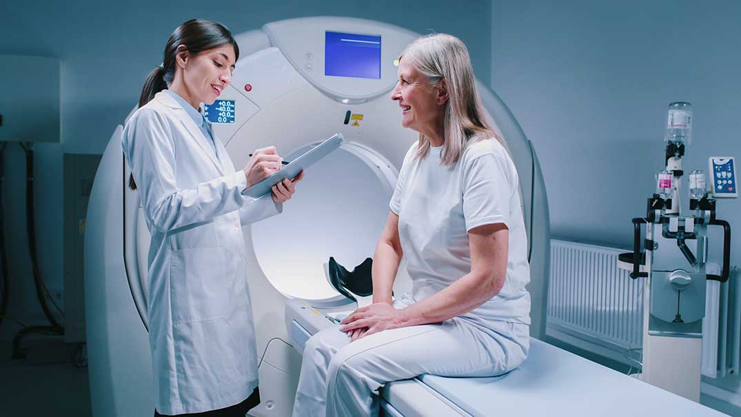 Frau sitzt vor MRT-Gerät und spricht mit einer Ärztin.